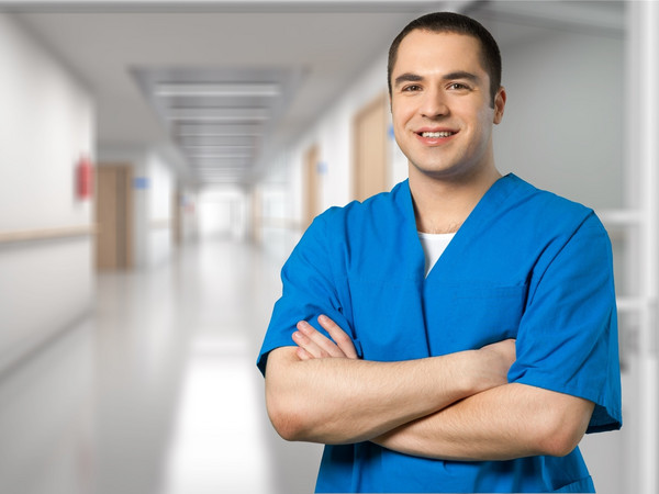 Ein junger Mann in blauer Pflegekleidung steht mit verschränkten Armen in einem hellen Krankenhausflur und lächelt freundlich in die Kamera. Der Hintergrund zeigt eine moderne, saubere Klinikumgebung mit Türen und Handläufen entlang des Gangs.